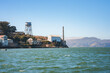 Scenic view of Alcatraz Island in San Francisco Bay, California, USA. Features former prison building, water tower, and coastal hills under blue sky. Peaceful waters.