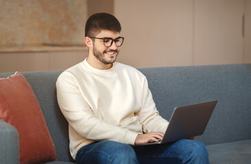 Aufkleber - A man wearing eyeglasses is sitting comfortably on a modern couch, engrossed in his work on a laptop computer, home interior