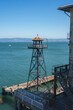 View from Alcatraz prison in San Francisco, USA. A guard tower stands on a pier with a weathered dome roof. A boat floats in the bay with a clear sky.