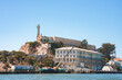 Sunny view of Alcatraz Island in San Francisco Bay. Former federal prison, main cellhouse, lighthouse, and rugged terrain depicted. Isolated beauty captured.