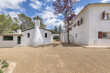 White facades of an Andalusian cortijo style country house with windows with red wooden shutters and terracotta and gravel floors on a clear sky day surrounded by leafy trees