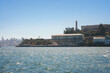 View of Alcatraz Island in San Francisco Bay, California, USA on a clear day. Iconic buildings and lighthouse against a blue sky, with cityscape in the background.