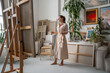 Thoughtful artist woman with inspiration paints picture, pondering ideas, holding in hands palette with oil paints in front of easel with canvas. Creative female standing barefoot in cozy art studio. 
