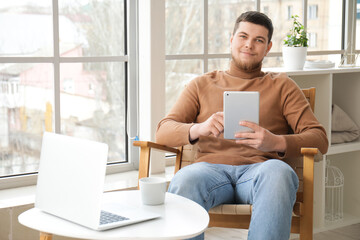 Poster - Young bearded man using tablet computer in armchair near window at home