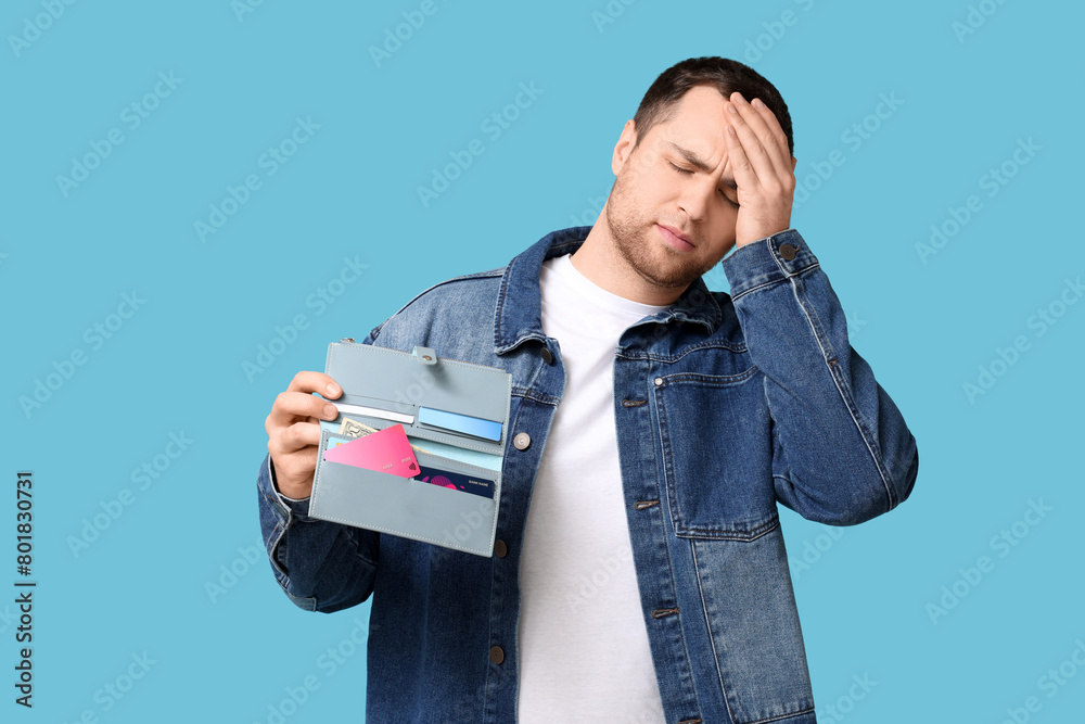 Young upset man holding purse with credit cards on blue background