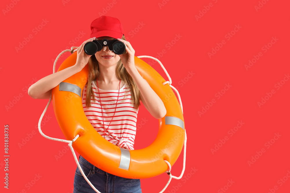 Young female lifeguard in cap with rescue ring looking in binocular on red background