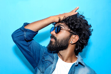 Young handsome indian man wearing blue shirt and sun glass standing over blue background with hand on head for stress.