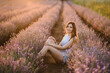 Smiling female sits among violet lavender flowers with sunlight on summer day. Woman in purple lavender field sniffing flowers at sunset. Young slim girl sitting and enjoying fun the time.