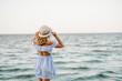 Young attractive blond woman in straw hat joyfully walks near sea. Portrait of stylish female closeup. Beautiful girl standing on beach ocean and enjoying sunny summer happy day on vacation. Back view