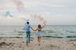 Happy married couple holds in hands smoke bomb. Baby shower. Twins: boy and girl. Back view. Man and woman having fun running on sand and holding colorful blue and red or pink smoke bombs on the beach