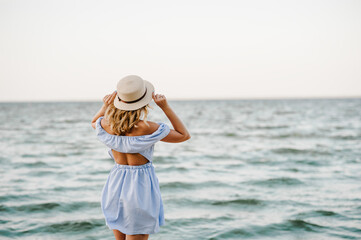 Young attractive blond woman in straw hat joyfully walks near sea. Portrait of stylish female closeup. Beautiful girl standing on beach ocean and enjoying sunny summer happy day on vacation. Back view