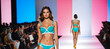 Elegant female models in turquoise swimwear confidently walking the runway at a summer fashion show event, with an audience in soft focus in the background