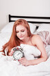 Sleepy and angry young beautiful woman holding an alarm clock lying on the bed. Insomnia problem.