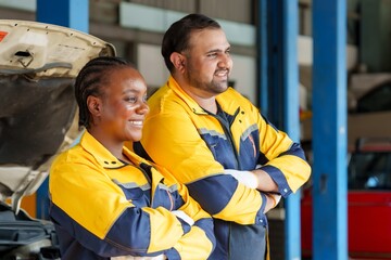 Two mechanics in yellow and blue uniforms, smiling confidently, arms crossed, standing in garage, with car and open hood in background, teamwork in automotive repair. Service work in car repair shop