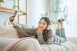 Smiling asian woman with headphones taking selfie with mobile smartphone on couch at home