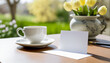 Springtime outdoor background, table with cup of tea and blank empty white cardboard card with copy space and spring flowers. Sunny day.