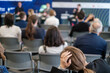 Focused image of a woman feeling stressed or overwhelmed while attending a business seminar. She sits among blurred audience members in a modern conference setting.