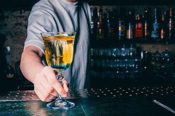 Wall Mural - person in grey attire holds a stemmed glass containing a yellowish cocktail. The individual stands behind a metal bar counter with circular holes, surrounded by various liquor bottles on shelves