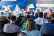 Focused audience members view a panel discussion at a conference. The image captures engagement and diverse professionals in a blurred background.