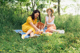 Fototapeta Miasto - A couple of women chatting while sitting on a blue picnic blanket in the open air