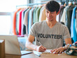 Male Charity Worker At Desk With Laptop Checking Clothing Donations At Thrift Store