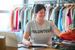 Female Charity Worker At Desk With Laptop Checking Clothing Donations At Thrift Store