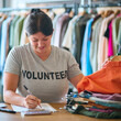 Male Charity Worker At Desk With Clipboard Checking Clothing Donations At Thrift Store