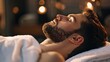 A bearded man lies back on a plush towel, his eyes closed in a serene spa setting with ambient warm lighting.