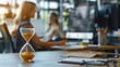 © ORG - Close-up of an hour glass with a woman sitting at a desk and working on a desktop computer in the background. Time and business concept