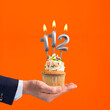 Hand holding birthday cupcake with number 112 candle - background orange