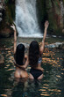 A couple of women in bikinis stand in front of a waterfall in Bali