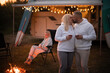 A married couple with glasses of wine stands against the background of a motorhome and rests together by the campfire. Evening family vacation