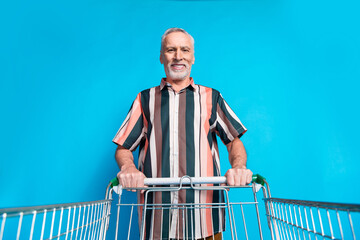 Sticker - Photo of smiling senior man wear summer striped stylish clothes hold trolley isolated on blue color background