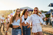 © artiemedvedev - Three young women walk together on sunny beach at music festival. Happy friends in trendy summer outfits enjoy live concert, dancing, laughing in festive atmosphere. Group of girls having fun by sea.