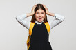 Surprised girl in school uniform and yellow rucksack keeping hands on head and staring at camera, isolated over white background. Back to school