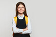 Delighted schoolgirl kid wearing uniform, eyeglasses and yellow backpack, holding crossed hands and smiling against white background, free empty space