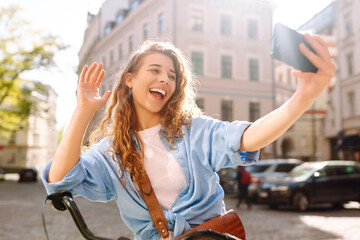 Young woman using mobile phone while  riding bicycle at the city street outdoor. Telecommunications, video call conference, communication with friends and family remotely. Selfie time.