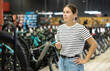 Interested thoughtful young female shopper casually walking through bicycle shop, looking over selection of modern bikes and gear available, contemplating best choice for cycling needs..