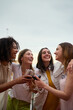 © CarlosBarquero - Vertical. Group multiracial smiling female friends toasting glasses red wine and celebrating party outdoors. Young funny women hugging cheers on rooftop. Happy girls enjoying summer vacation evening