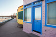 Pastel building with lavender exterior, blue door and window frames marked 6 1 2 . Yellow and green boutique. Pier in background, likely near water. Sunny, quiet street view.