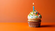 Tasty cupcake with blue candle and rainbow sparkle on orange background