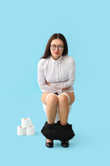 Poster - Young Asian businesswoman with bellyache sitting on toilet bowl against blue background