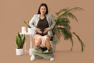 Poster - Young Asian woman with cramps sitting on toilet bowl against beige background