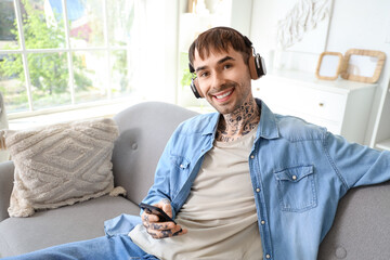 Wall Mural - Young tattooed man in headphones with mobile phone listening to music on sofa at home