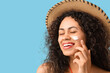 Young African-American woman applying sunscreen cream on her face against blue background, closeup
