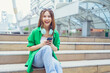 Young Asian woman using smartphone enjoying music listening through wireless headphones while sitting on steps outside in the city.