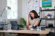© Wasana - A woman is sitting at a desk with a laptop and a cell phone. She is talking on the phone and she is in a serious mood