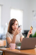 © Wasana - A woman is sitting at a desk with a laptop and a cell phone. She is looking at her cell phone and holding a credit card. Concept of modern technology and convenience