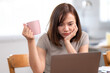 © Wasana - A woman is sitting at a table with a laptop and a pink cup. She is looking at the laptop with a frown on her face