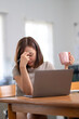 © Wasana - A woman is sitting at a table with a laptop and a pink mug. She is looking at the laptop and she is in a state of distress